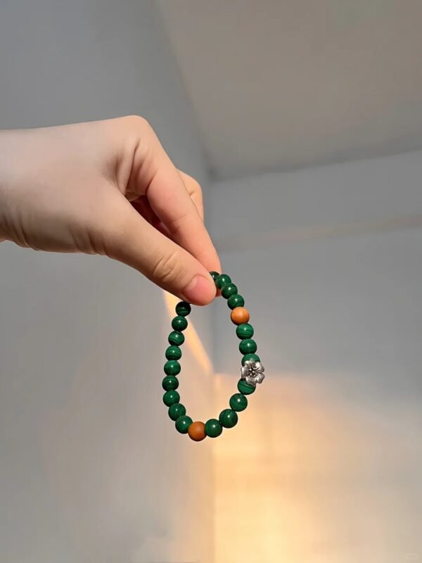 Holding single-strand malachite bracelet with red agate spacers and silver flower charm, wood-fire-metal energy blooms